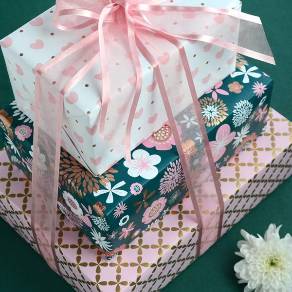 A stack of 3 boxes, gift wrapped in hearts, floral and geometric designs. Finished with a satin edge sheer pink ribbon